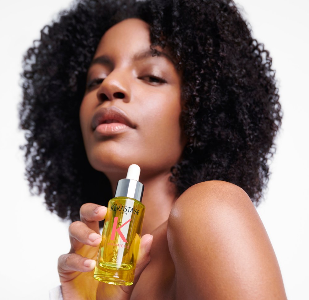 Ebonee Davis, avec ses superbes cheveux bouclés, tient la meilleure huile capillaire pour cheveux abimés : Première Huile Gloss Réparatrice.